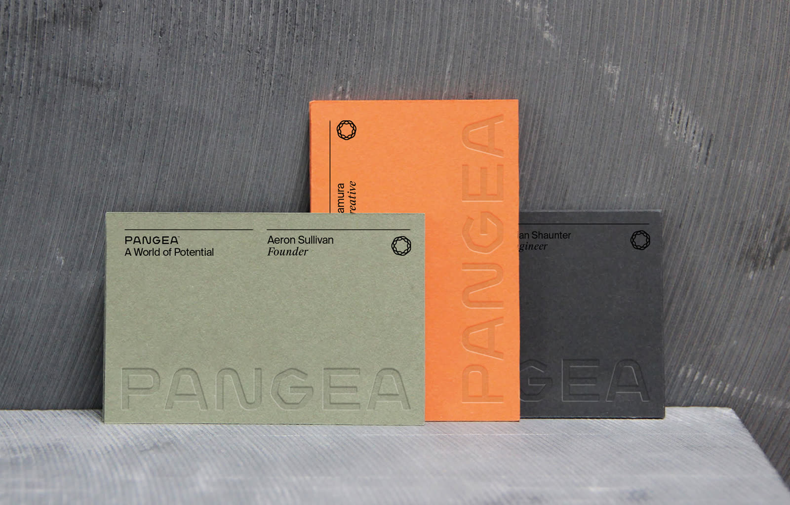 Three Pangea business cards in sage green, orange, and black leaning against each other on a textured concrete surface, each embossed with the Pangea wordmark.
