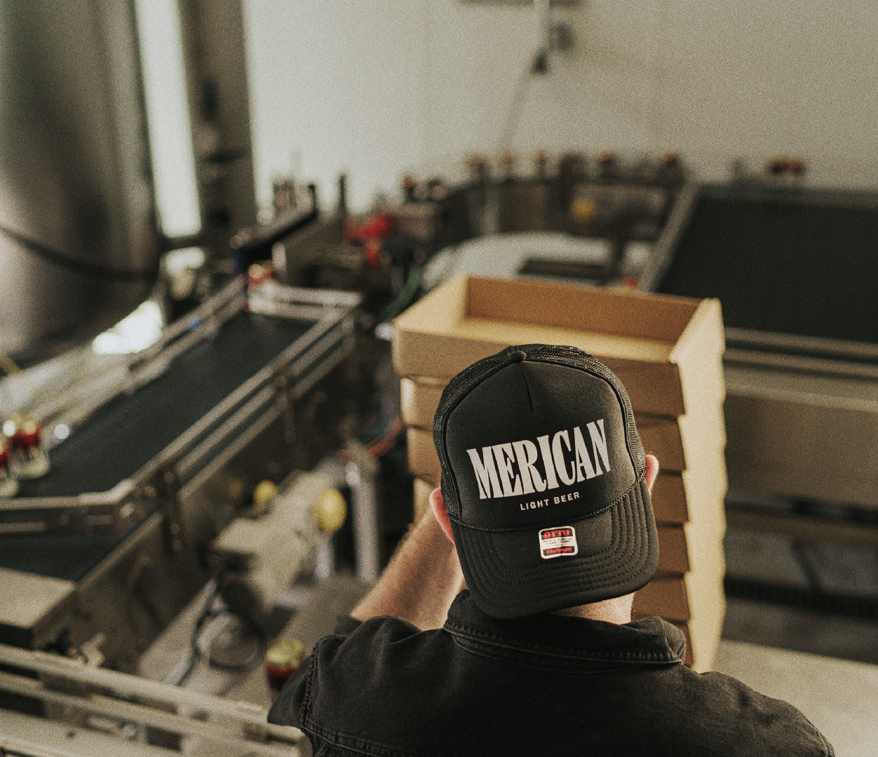 Merican staff member packing cans into cardboard boxes, wearing a backwards black trucker cap with the Merican logo printed on the front.