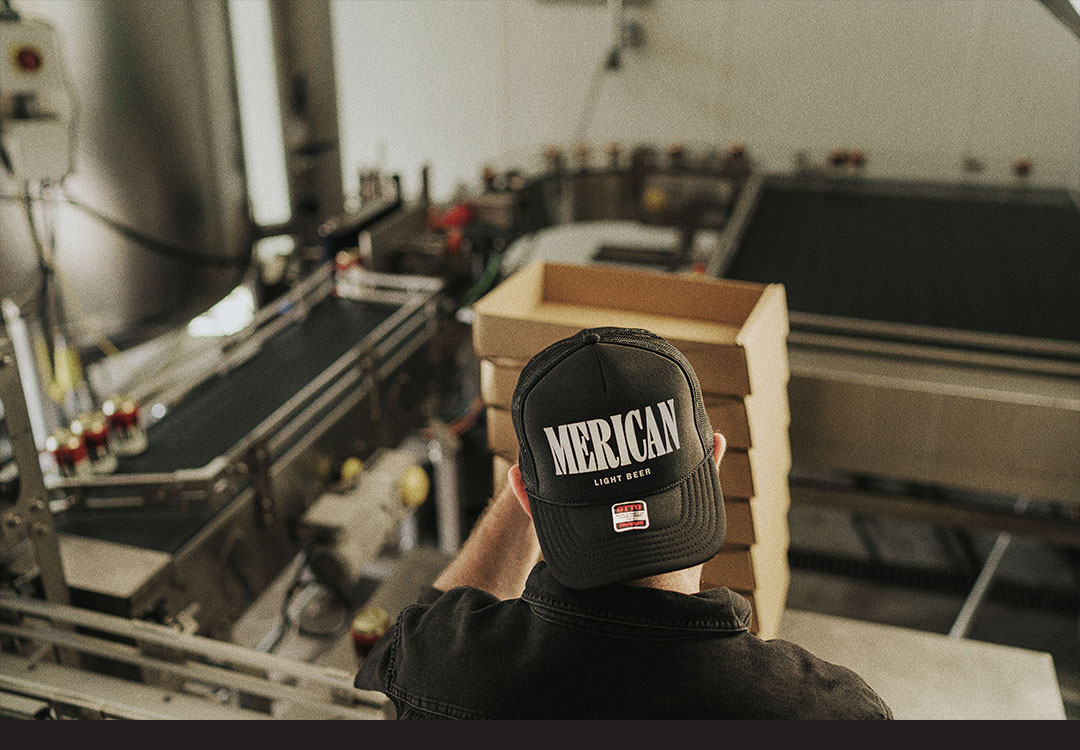 Merican staff member packing cans into cardboard boxes, wearing a backwards black trucker cap with the Merican logo printed on the front.
