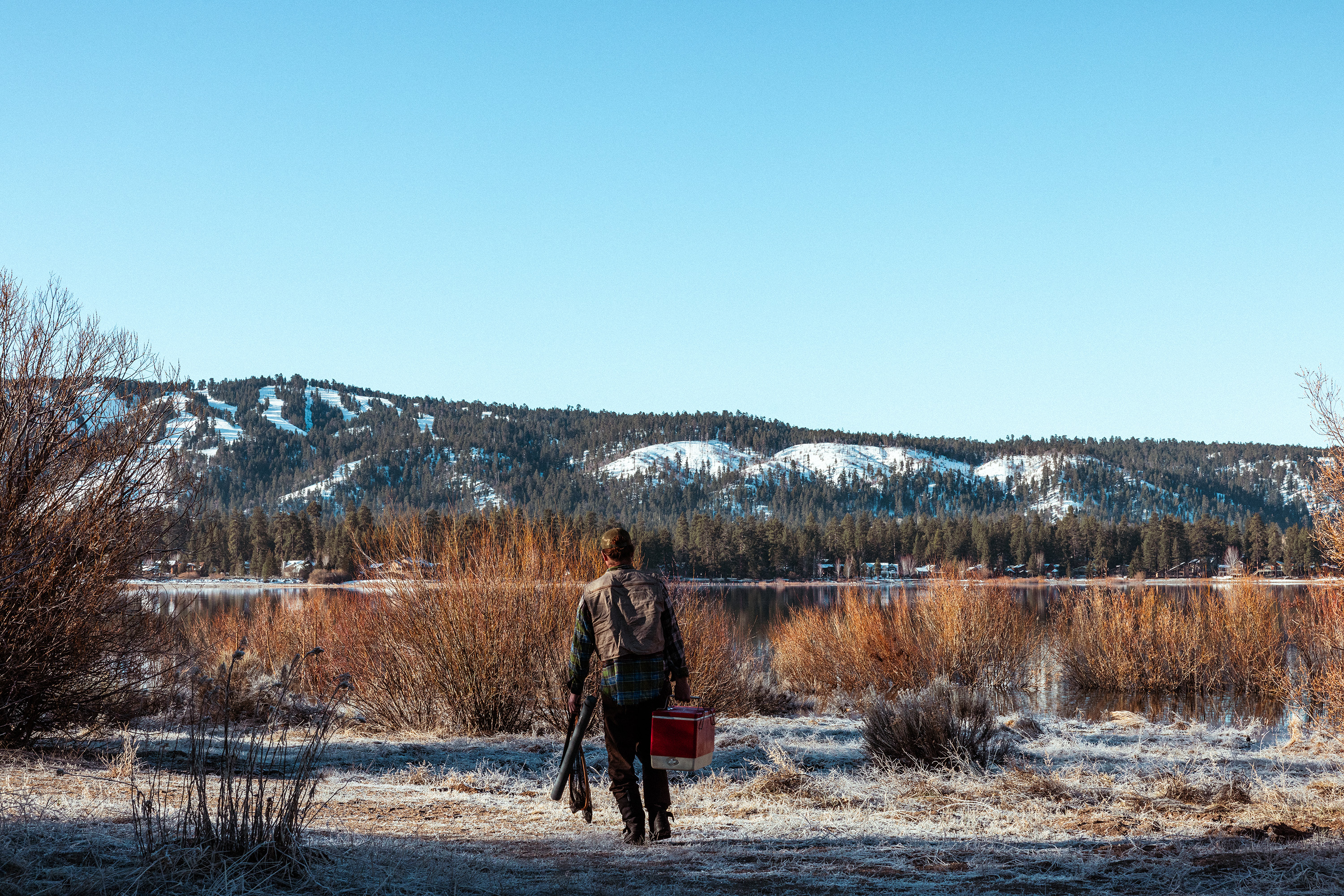 Man with his back turned, carrying a fishing rod in one hand and a red cooler in the other, admires the winter landscape and lake in front of him.