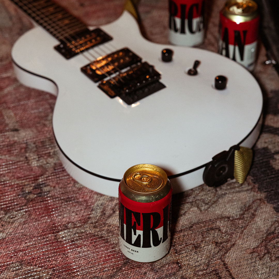 White electric guitar laid on a faded rug, surrounded by cans of Merican beer.