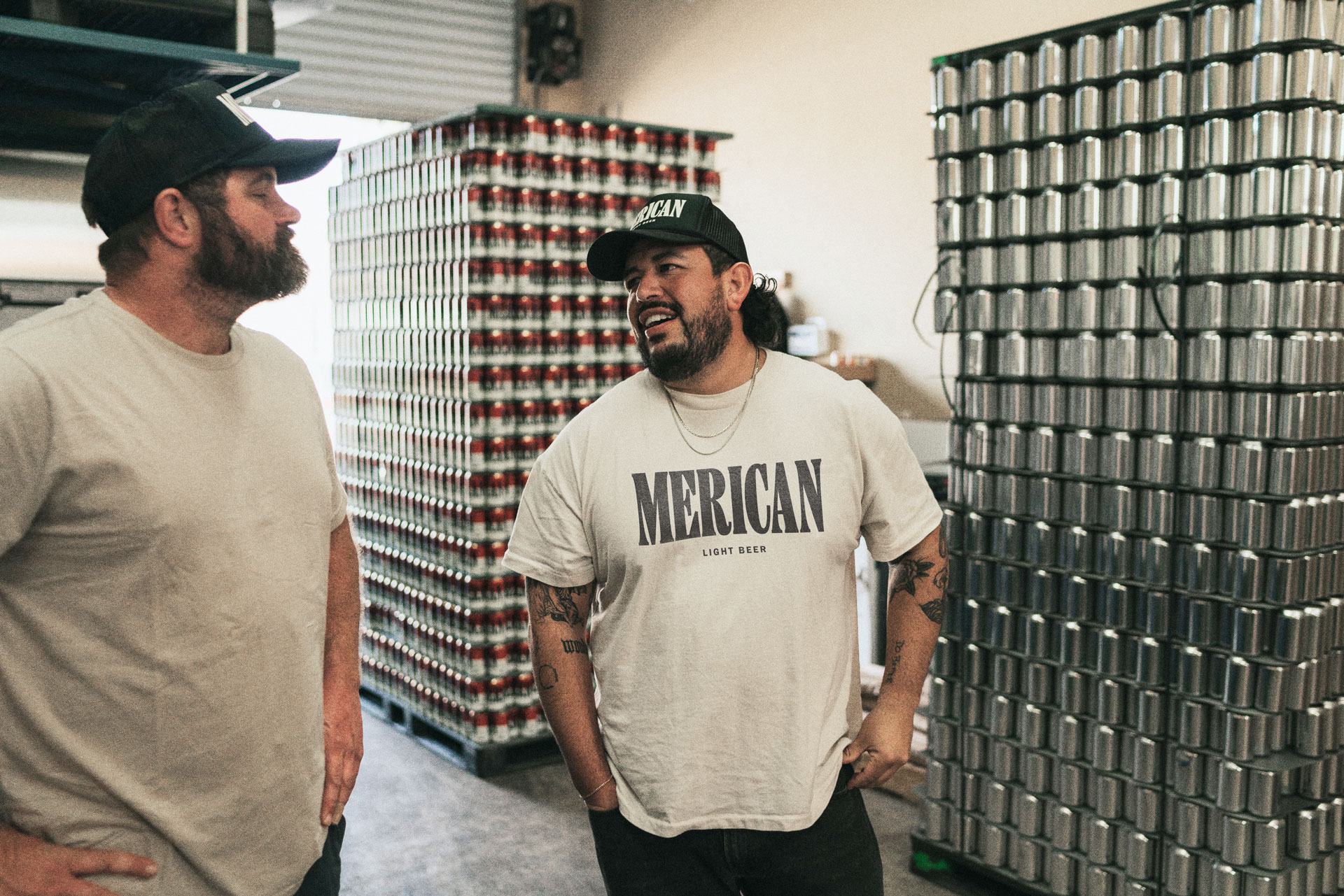 Two Merican staff members talking to each other in the production facility, with large stacks of aluminium beer cans behind them.