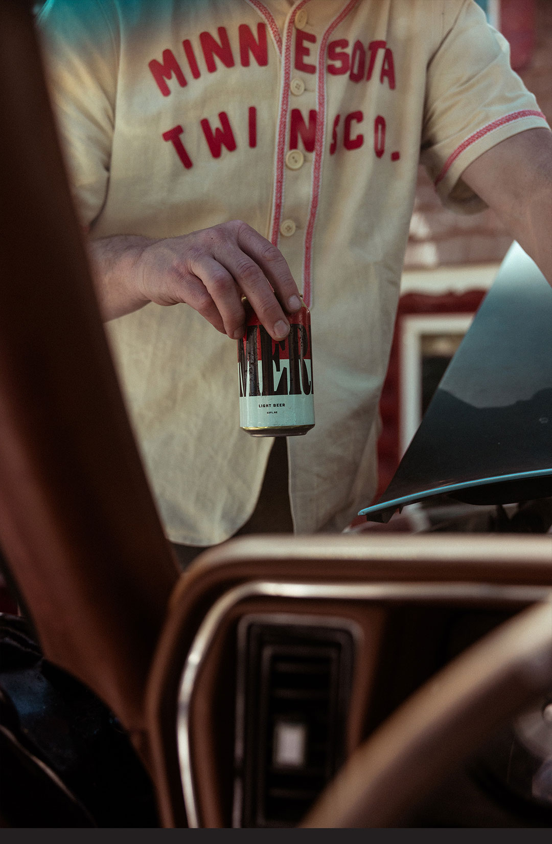 White man outside, pictured through the windscreen of a vintage car, holding a can of Merican beer, wearing a cream-colored Minnesota Twins shirt.